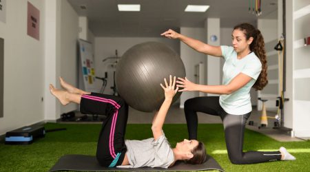 Physical therapist assisting young caucasian woman with exercise with yoga ball during rehabilitation in the gym at hospital. Female physiotherapist training a patient in physiotherapy center.